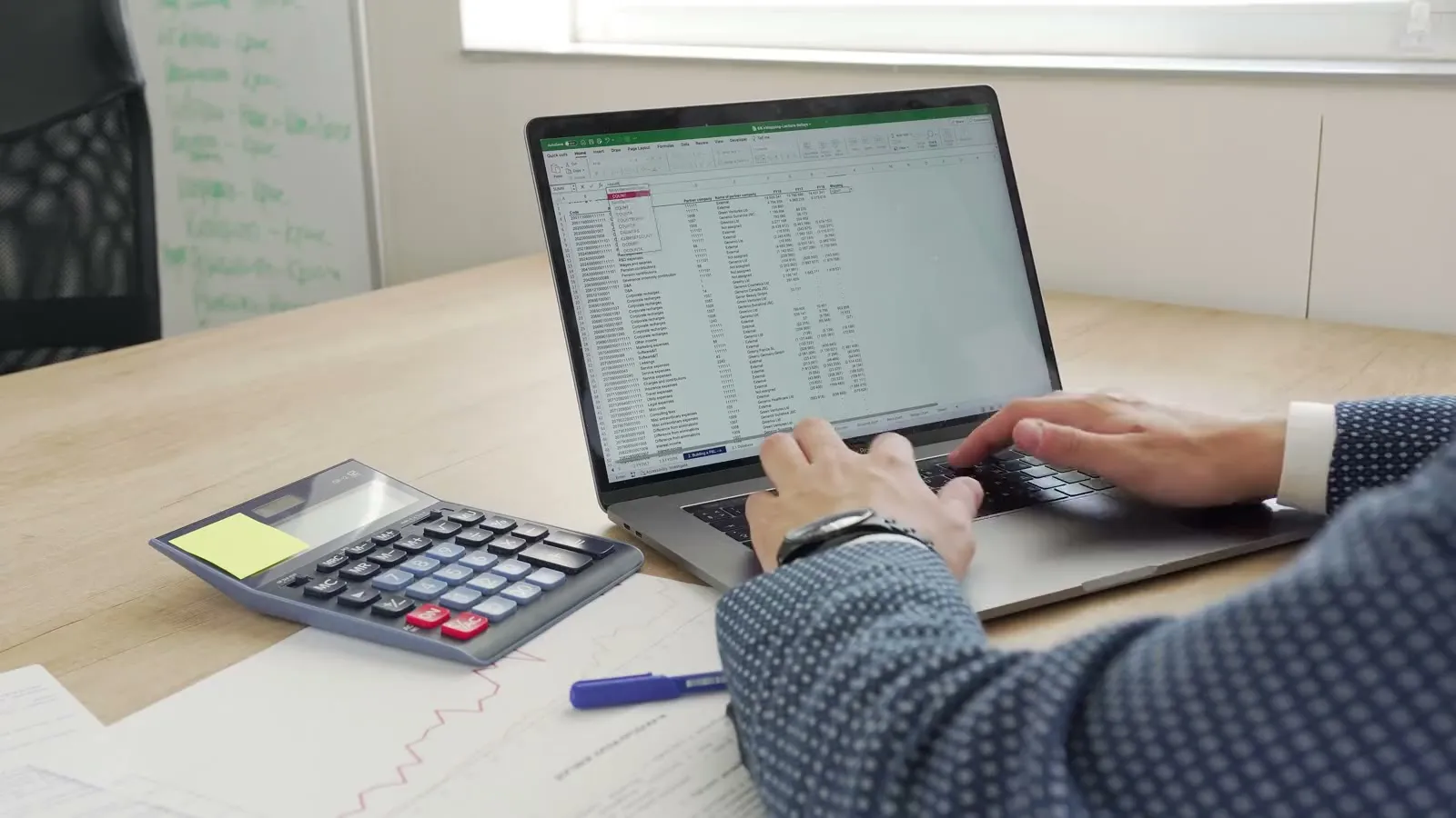 Hands working through a spreadsheet on a laptop, beside a calculator and printed reports